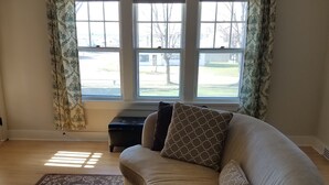 Un salón con un sofá color beige, cojines estampados, un reposapiés redondo y una alfombra con diseño. Hay tres ventanas con cortinas y una vista a un patio exterior.
