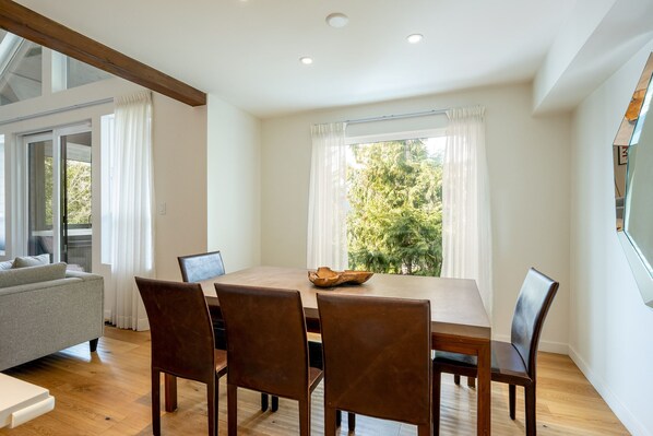 Un comedor con una mesa de madera, cuatro sillas y un ordenador portátil. Hay un sofá y una ventana con cortinas.