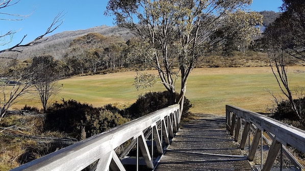 A wooden bridge leading to a grassy field with trees and hills in the background. - CHILL-OUT - Thredbo Village - moments to the mountain. Winter and summer! (Thredbo Village)