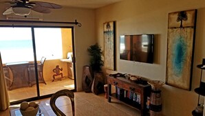 Ein Wohnzimmer mit einem großen Fernseher, einer Holzeinrichtung mit Büchern, einer dekorativen Schwanenfigur und Blick auf den Strand durch eine Schiebetür.
