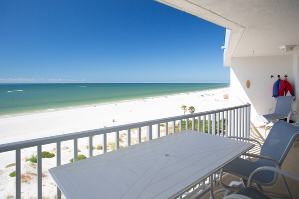 A balcony with a white table and chairs, overlooking a beach with clear blue water and white sand.