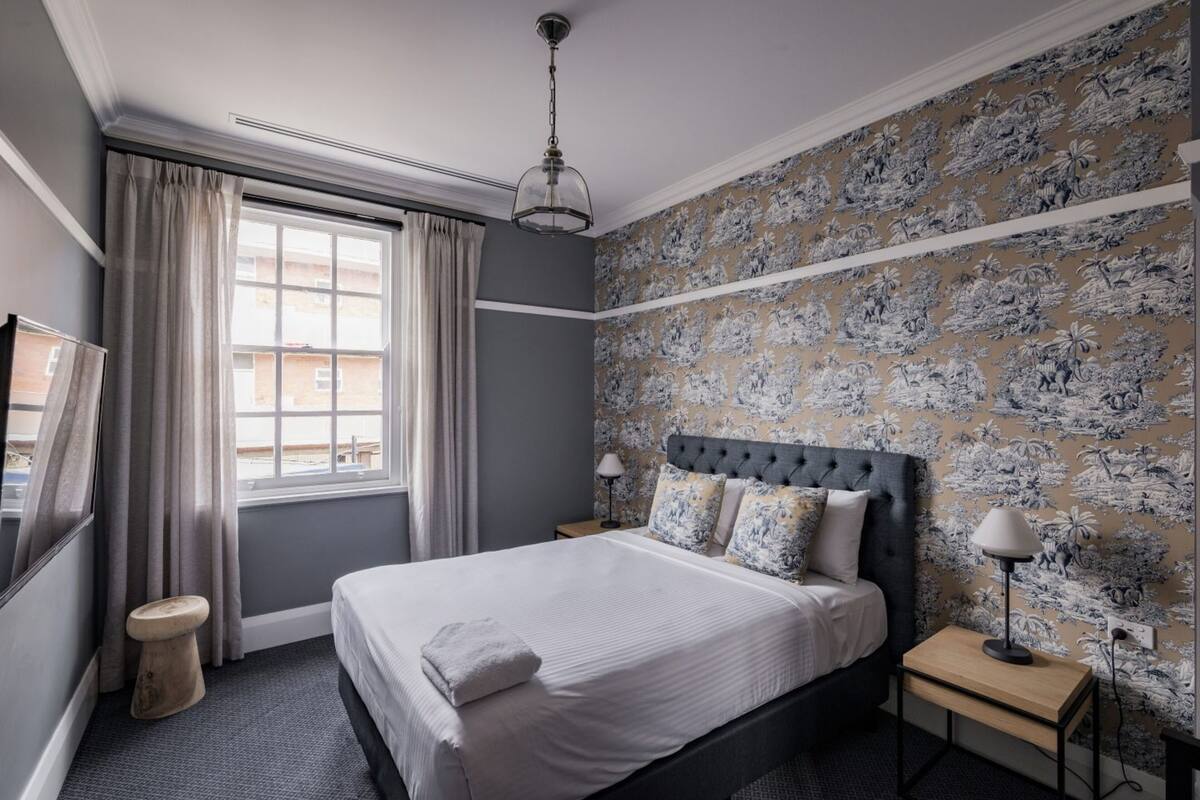 A bedroom with a bed, a nightstand, a lamp, a window with curtains, and a wall with a floral pattern.