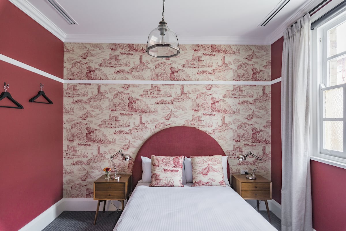 A bedroom with a red and white patterned wallpaper, a large bed with white bedding, two bedside tables with lamps, and a hanging light fixture.