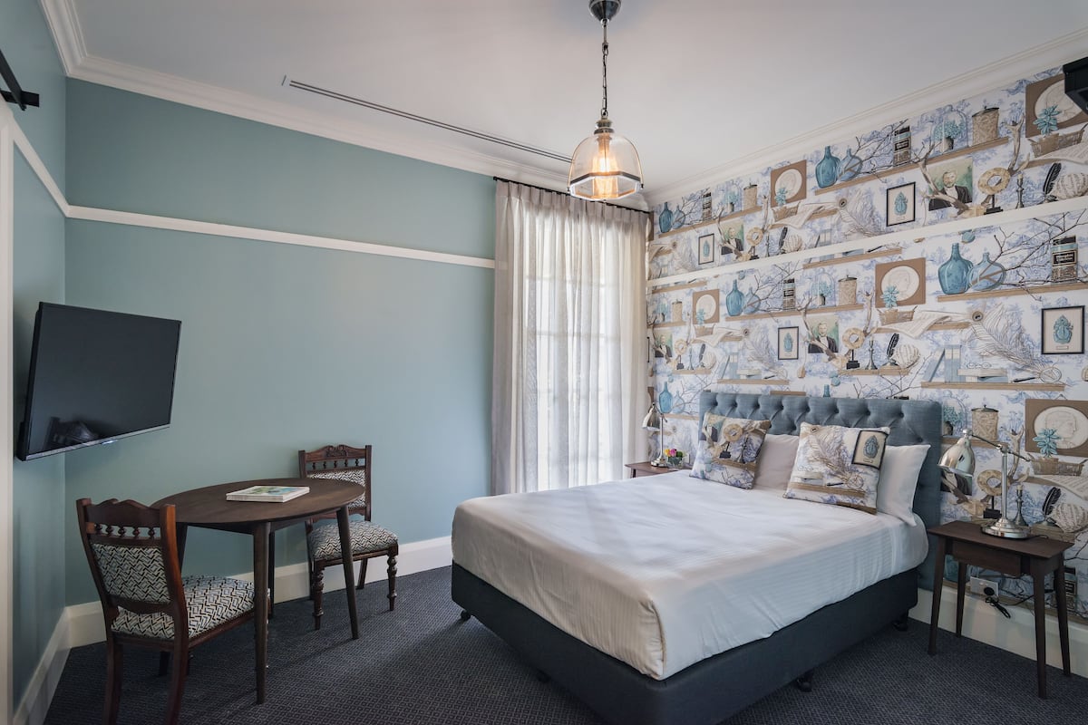A bedroom with a bed, a desk, chairs, a TV, and a patterned wallpaper.