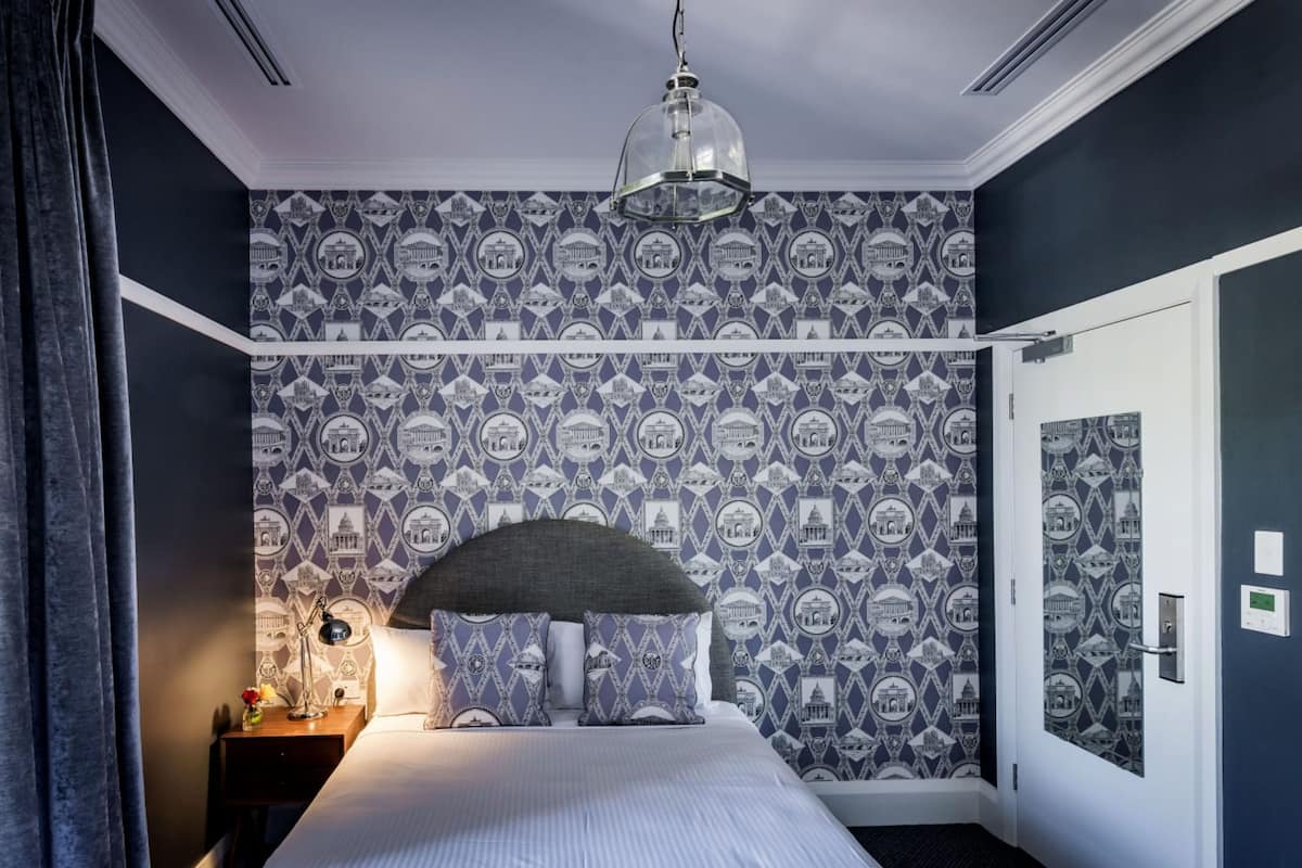 A bedroom with a patterned wallpaper, a bed with a grey headboard, a bedside lamp, and a hanging light fixture.