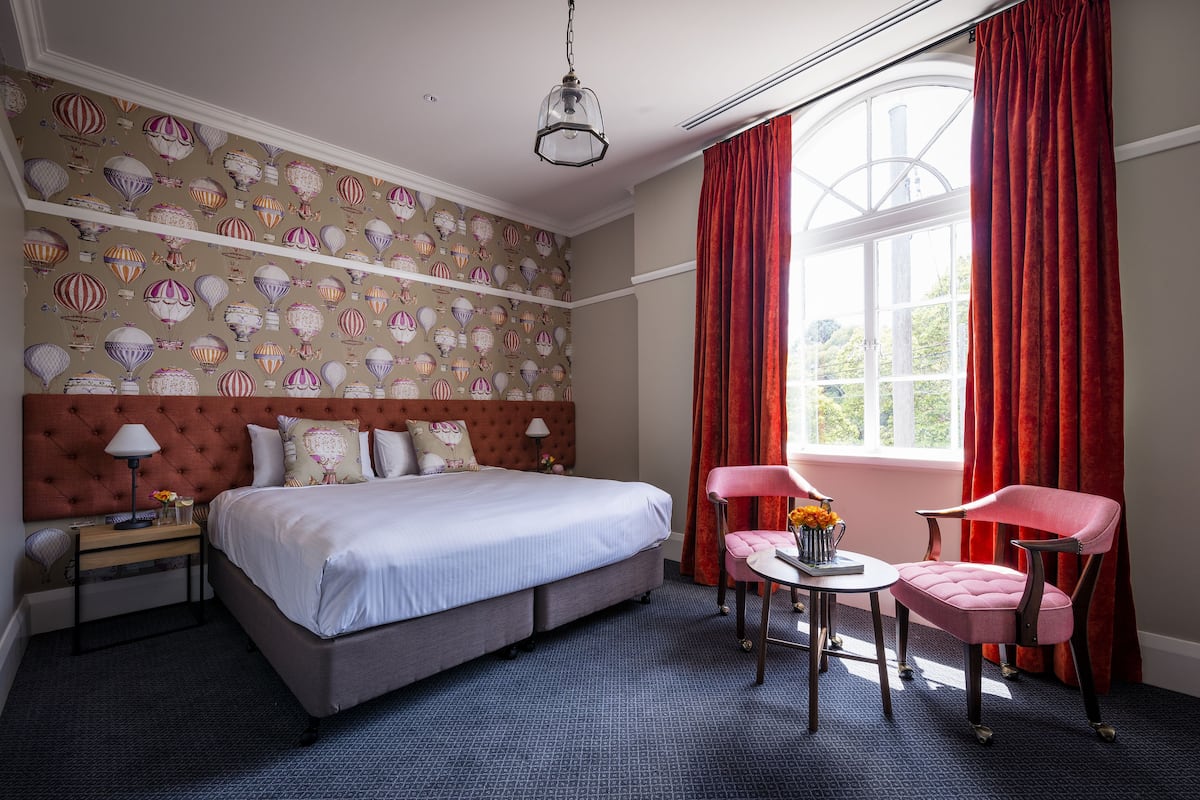 A hotel room with a large bed, a nightstand, a desk with a chair, and a window with red curtains.