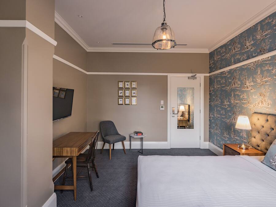 A hotel room with a bed, a desk, a chair, a small table, a lamp, and a ceiling fan.