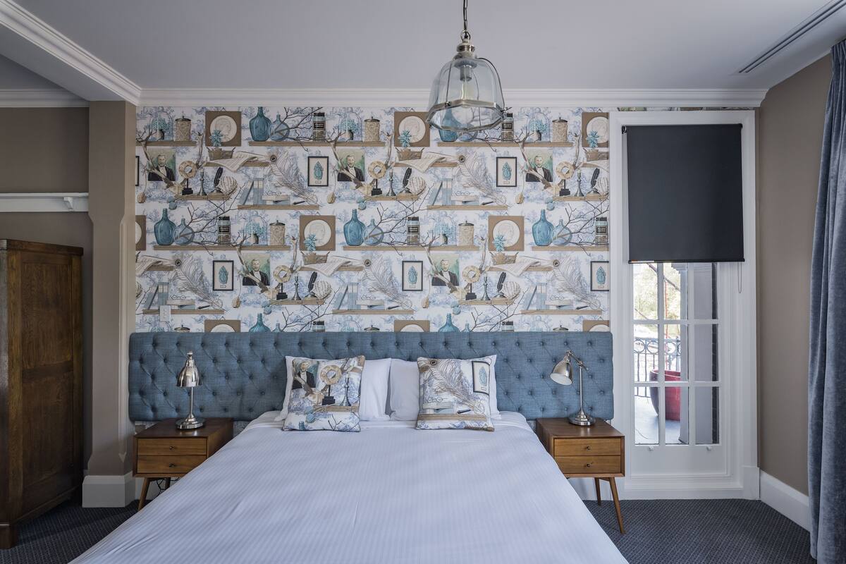 A bedroom with a large bed, a tufted headboard, a patterned wallpaper, a hanging light fixture, and a window with a view.