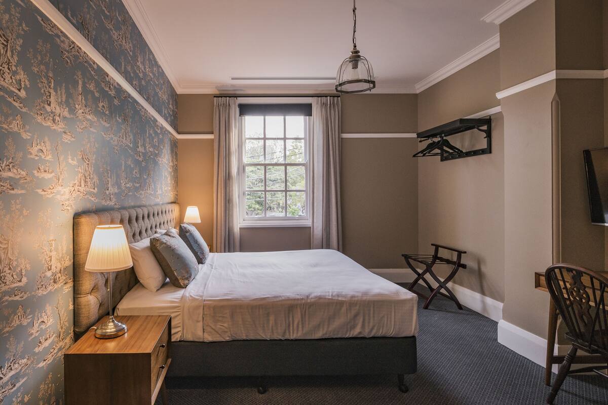 A hotel room with a large bed, a bedside table with a lamp, a window with curtains, a chair, and a coat rack.
