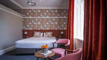 A hotel room with a bed, a chair, a table with a vase of flowers, and a patterned wallpaper.