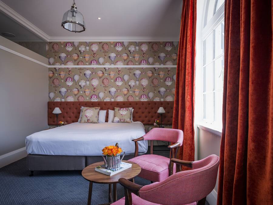 A hotel room with a bed, a chair, a table with a vase of flowers, and a patterned wallpaper.