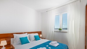 A hotel room with a wooden headboard, white and blue bedding, a window with a view, and a small plant on the windowsill.