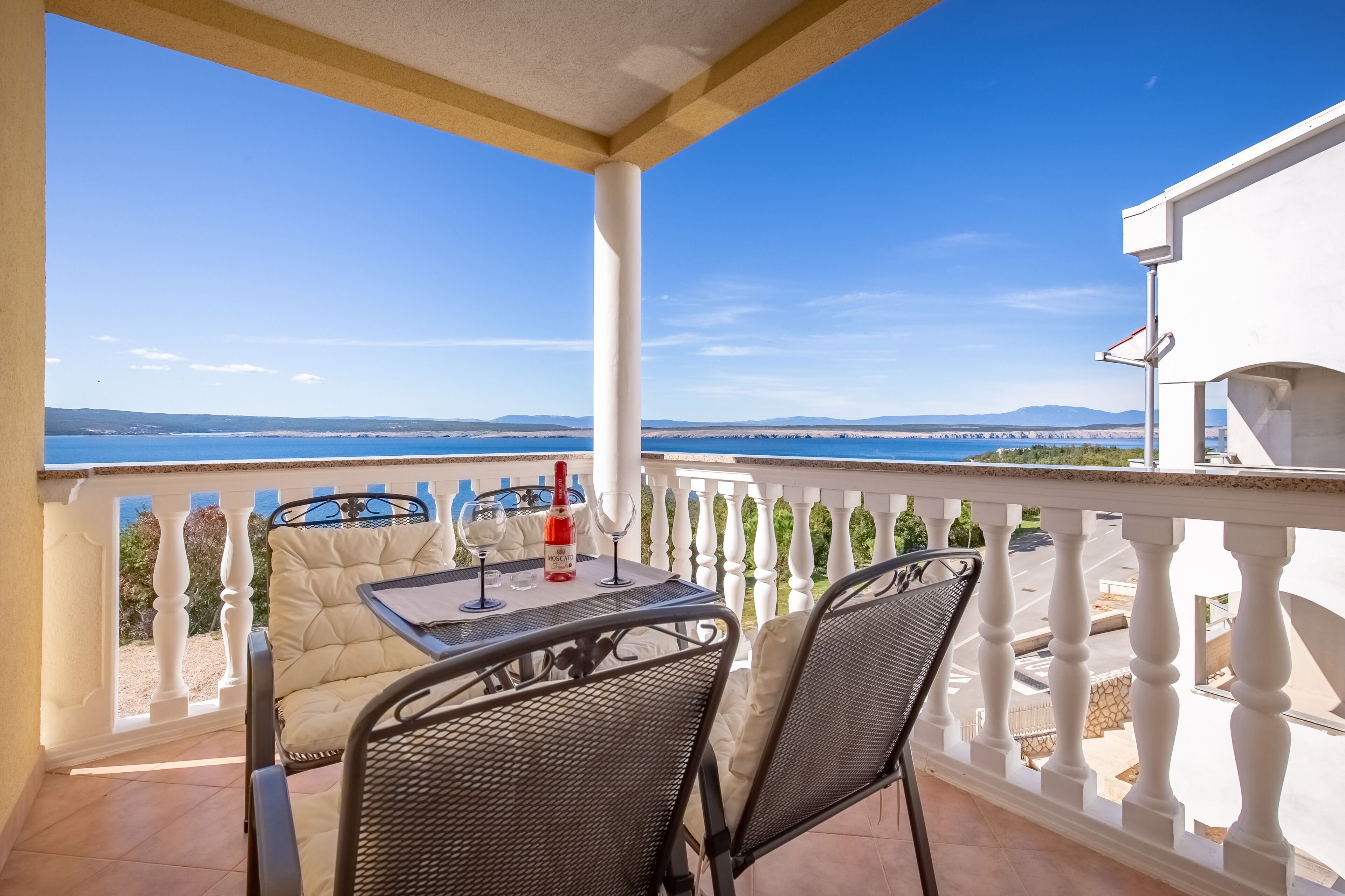 A balcony with a table and chairs, a bottle of wine, and a view of the sea and distant mountains.