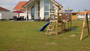 En legeplads med rutsjebane og klatrestruktur, en rød parasol og en hvid bygning med rødt tag i baggrunden.