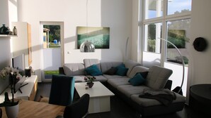 En moderne stue med sofa, sofabord og spiseplads med bord og stole. Der er et billede på væggen, og et vindue giver naturligt lys.