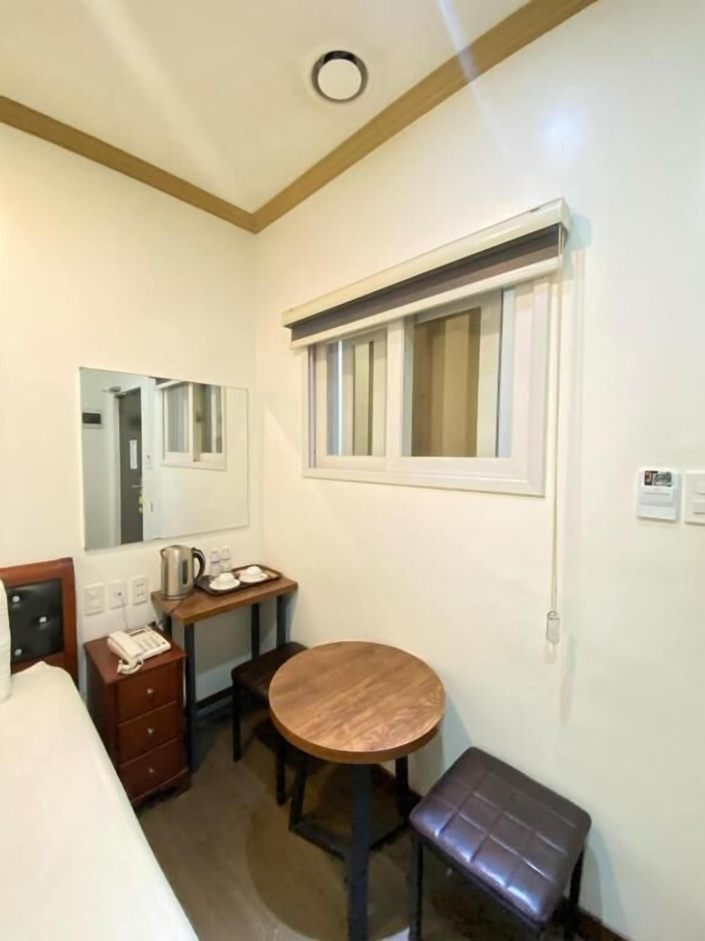 Een kleine, compacte hotelkamer met een enkel bed, een nachtkastje met telefoon, een klein rond tafeltje met een bankje, een raam met jaloezieën en een spiegel aan de muur.
