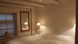 A bedroom with a large bed, a window with blinds, a mirror, and two wall-mounted lamps. - Romantic Frisian house on the East Frisian coast (Krummhörn)