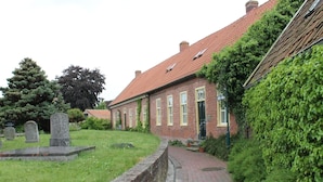 A brick building with a red-tiled roof, a brick pathway leading to the entrance, and a grassy area with headstones. - Romantic Frisian house on the East Frisian coast (Krummhörn)