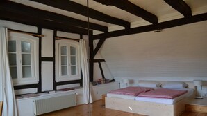 A bedroom with a sloped ceiling, wooden beams, a bed with pillows, a bedside table with a lamp, and a window with curtains. - Idyllic half-timbered house with a large garden and playground and orchard (Cleebourg)