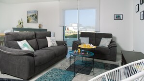 Uma sala de estar moderna com dois sofás cinzas, uma mesa de centro de vidro e uma cadeira branca. Há vista para o exterior através de uma grande janela.