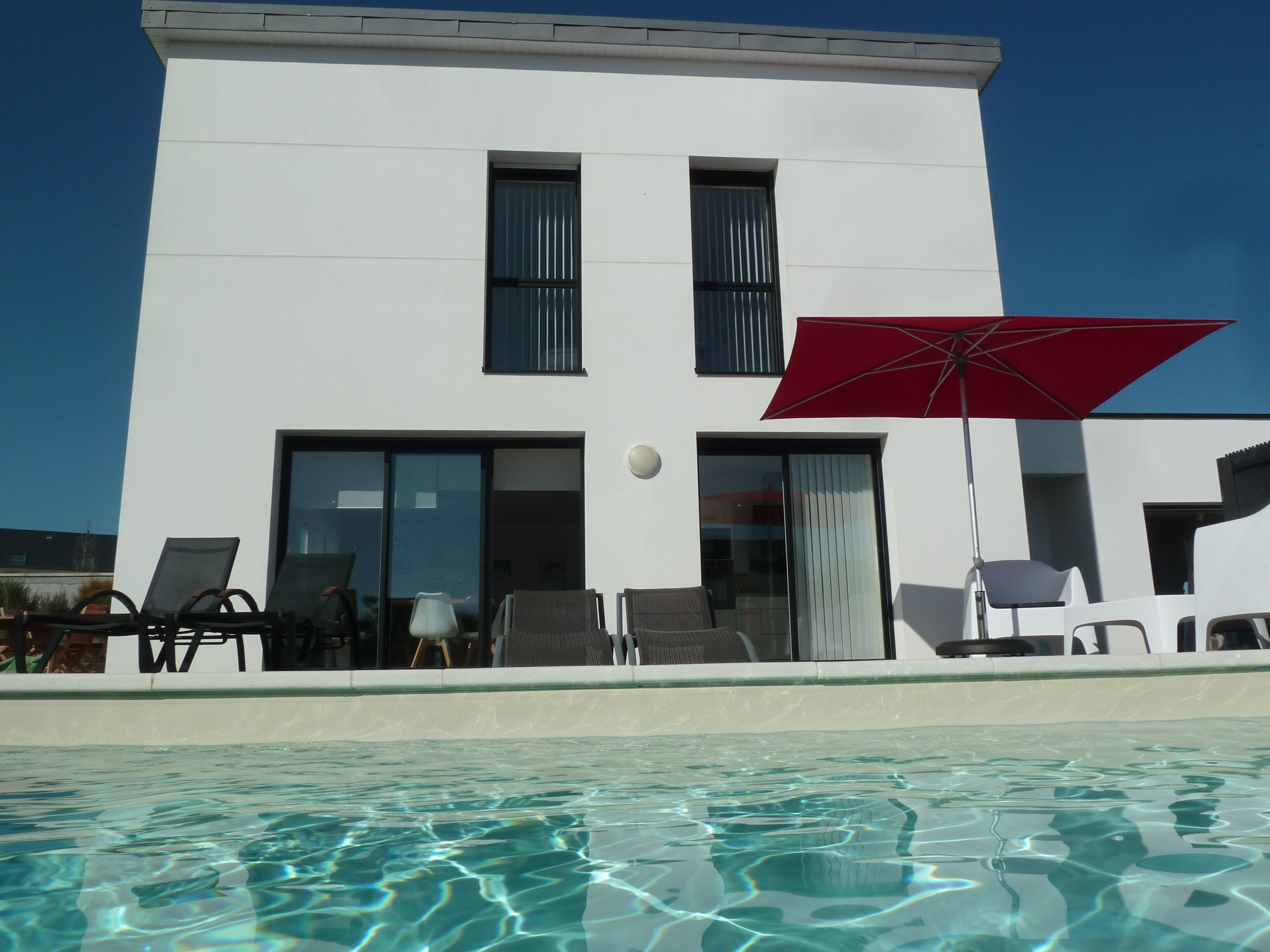 A modern building with a pool, featuring a red umbrella, lounge chairs, and a clear blue sky.