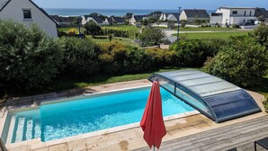 Piscina com cobertura de vidro, cercada por um deck de madeira e um guarda-sol vermelho.