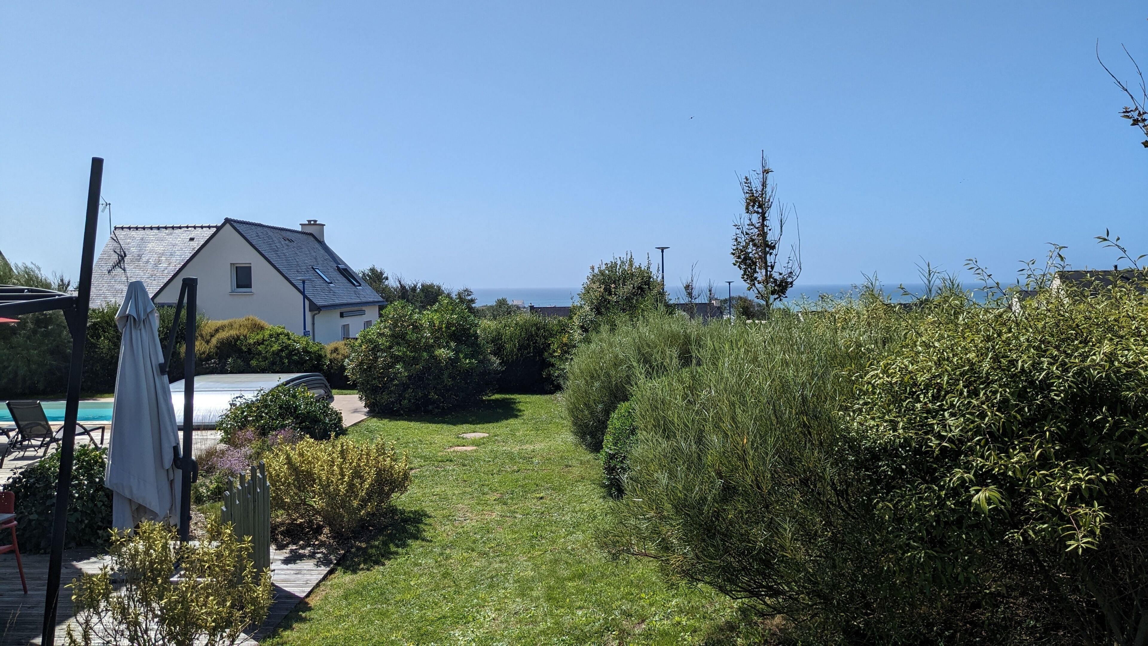 A house with a garden path leading to a pool, surrounded by greenery and a clear view of the sea.