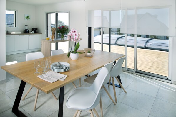 A modern dining area with a wooden table, white chairs, and a view through large windows.