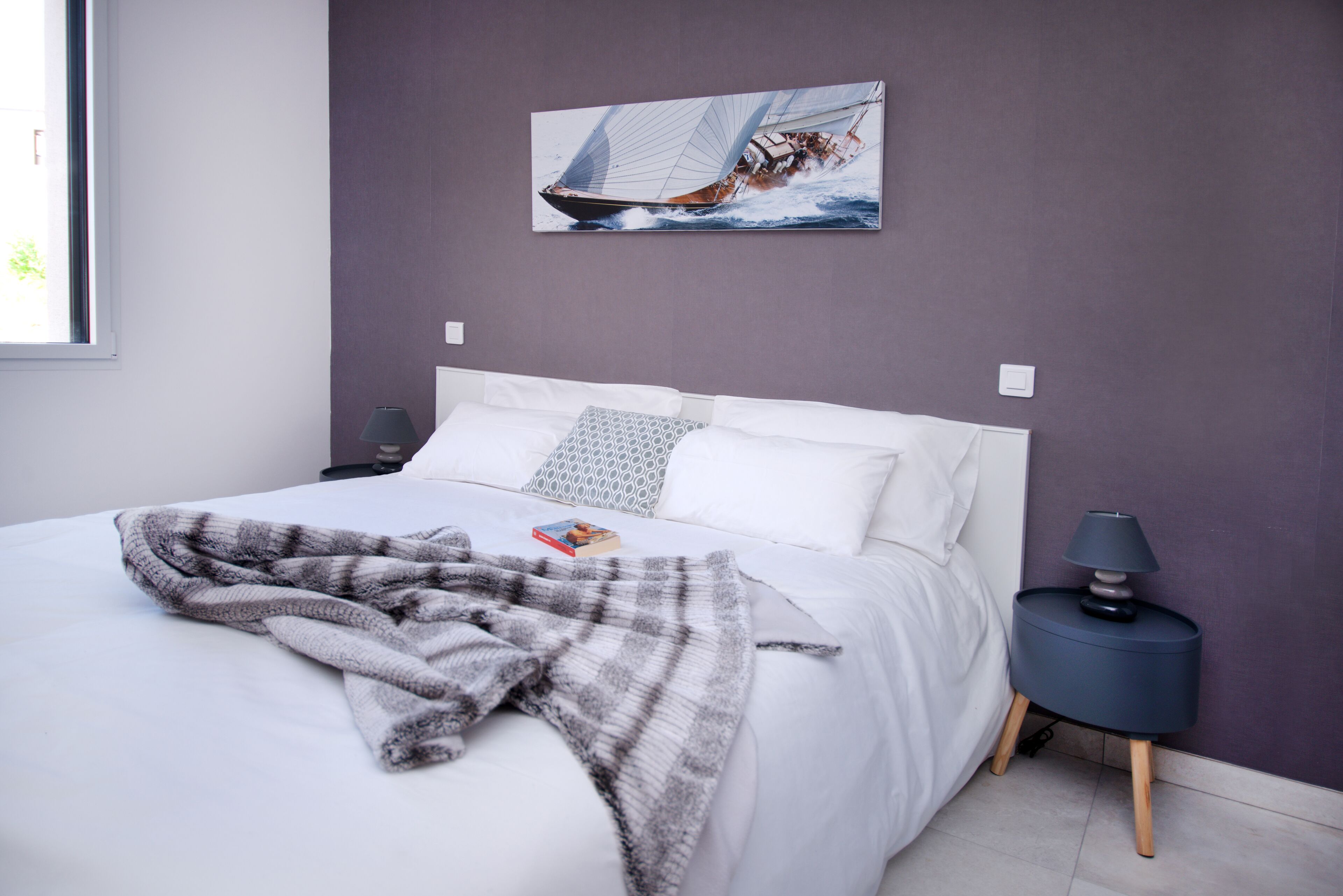 A neatly made bed with a gray and white striped blanket, a sailboat painting on the wall, and a small bedside table with a lamp.