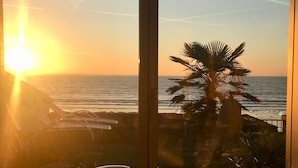 Vista a través de un cristal que muestra una zona de playa con una palmera, sillas de descanso y un coche estacionado afuera.