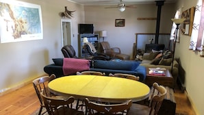 Un salón acogedor con una mesa redonda amarilla rodeada de sillas de madera, un sofá azul y un sillón de cuero marrón. Un televisor está montado en la pared, y se exhibe un mapa de Montana.