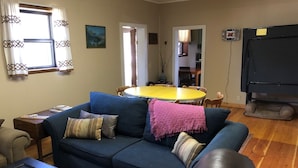 Una sala de estar con un sofá azul, una mesa de centro y una mesa de comedor con sillas. Se ve una ventana con cortinas, un cuadro en la pared y un televisor sobre un mueble.
