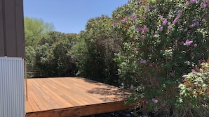 Una terraza de madera con base de piedra, rodeada de follaje denso y plantas florecientes.