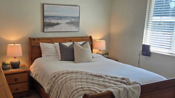 A bedroom with a wooden bed, two bedside lamps, a nightstand, a mirror, a wall-mounted painting, and a window with blinds. - The Suite Spot is a lovely two bedroom suite in a quiet neighborhood (Victoria)