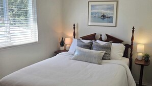 Una cama bien hecha con cabecera, dos mesitas de noche con lámparas, un cuadro en la pared y una ventana con persianas.