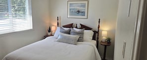 A neatly made bed with a headboard, two bedside tables with lamps, a framed picture on the wall, and a window with blinds.