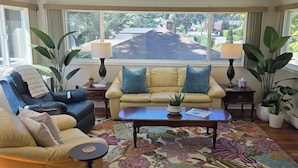 A sunlit room with a sofa, armchair, and coffee table, large windows with curtains, and potted plants. - The Suite Spot is a lovely two bedroom suite in a quiet neighborhood (Victoria)