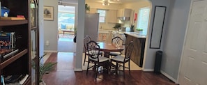 A dining area with a wooden table and chairs, a kitchen in the background, and a bookshelf with books.
