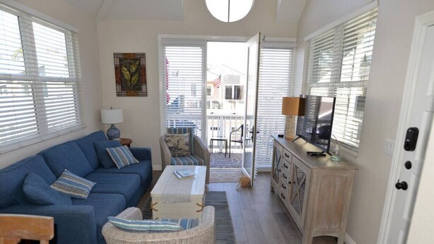 Un salon avec un canapé bleu, un fauteuil en osier, une table basse et un téléviseur posé sur un meuble en bois. Une porte donne sur un espace extérieur d’où l’on peut voir une maison et un drapeau.