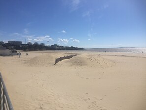 A sandy beach with a wooden walkway leading to the water. There are buildings in the background and a clear sky.