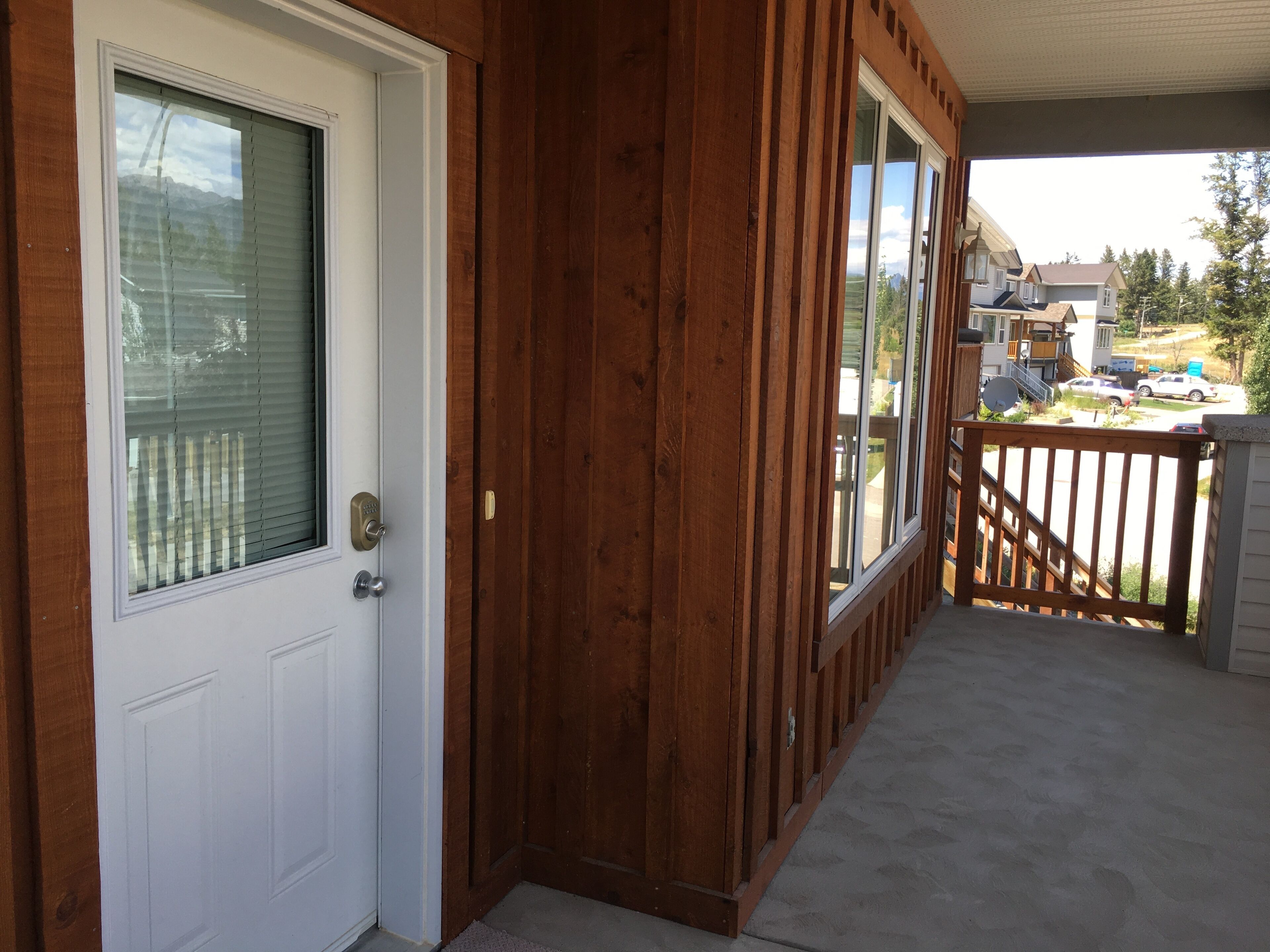 Une porte d’entrée en bois avec une fenêtre, un porche doté d’une balustrade en bois et une vue sur une rue résidentielle.