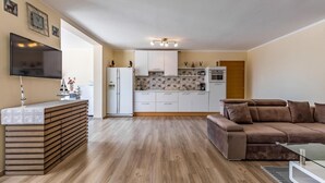 Una cuina moderna amb armaris blancs, un forn de sobre i un refrigerador. Hi ha una taula de menjador de fusta amb un mantell blanc i un objecte decoratiu a sobre. Un sofà marró es troba a la zona d'estar, i una televisió està penjada a la paret.