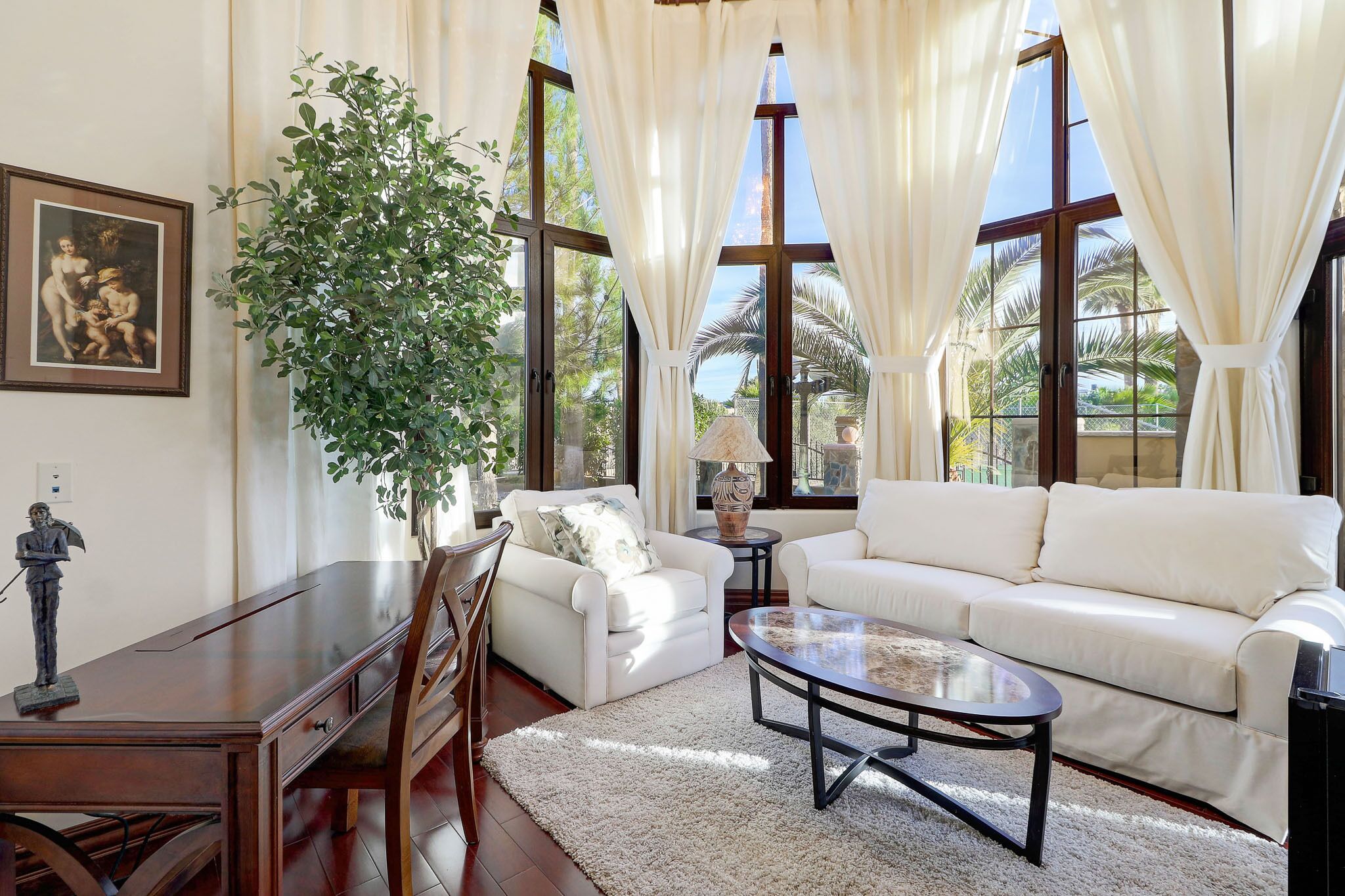 A spacious living room with a white sofa, a coffee table, and a dining table. There is a large plant and a statue on the wall.