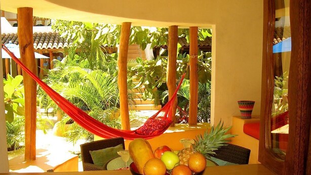 A room with a wooden table, a bowl of fruit, a hammock, and a view of a lush garden. - Beautiful Pool Side Condo, Ground Level.  Close to Beach & Town Center   (zihuatanejo)