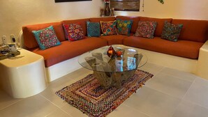 A living room with an orange sectional sofa, colorful pillows, a glass coffee table, and a woven rug. - Beautiful Pool Side Condo, Ground Level.  Close to Beach & Town Center   (zihuatanejo)