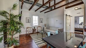 A spacious living area with a wooden floor, a dining table, and a large potted plant. There is a sofa and a window with blinds. - Perfect 2bd Suite w. Porch, Near King Street! (Charleston)