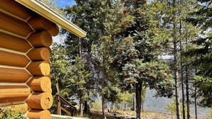 A log cabin with a deck overlooking a lake, surrounded by trees and a grassy area. - Rustic Waterfront Log Cabin (Kaslo)