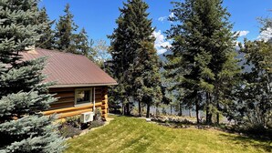 A log cabin with a metal roof, surrounded by dense evergreen trees and a clear blue sky. - Rustic Waterfront Log Cabin (Kaslo)