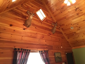 Interior de cabana de madeira com duas cabeças de veado penduradas no teto, um claraboia e um quadro na parede.