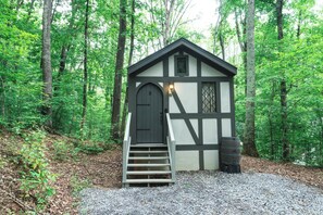 A small, single-story cabin with a dark door and a set of stairs leading up to it, surrounded by a forest.